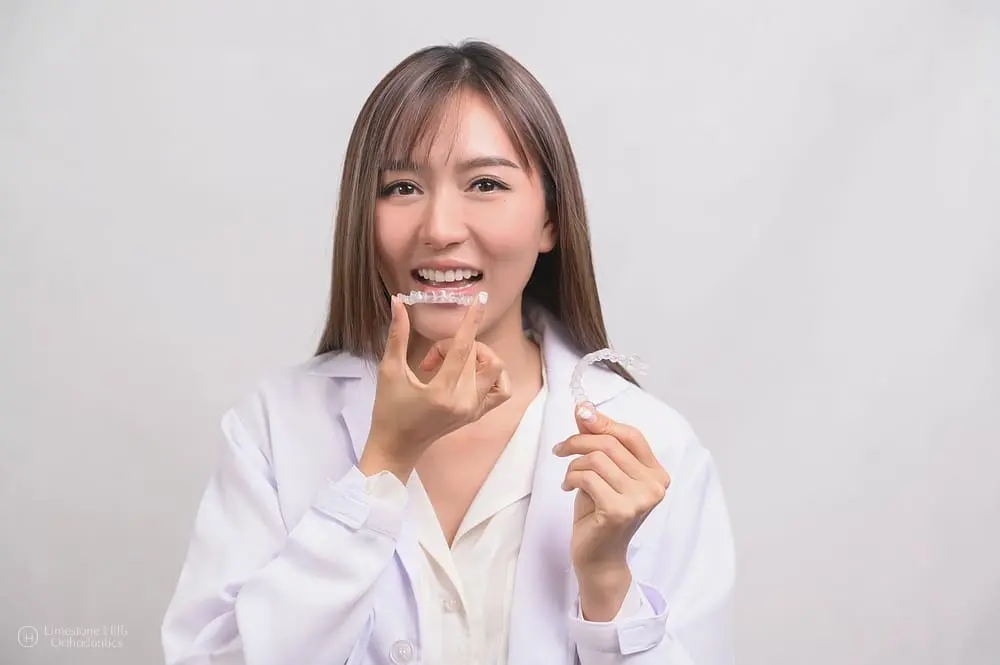 A woman in a white coat inserts a clear aligner at Limestone Hills Orthodontics in Austin, TX, against a plain background.