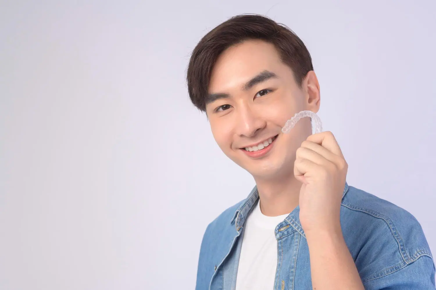 Smiling young man in a denim shirt holds a clear dental aligner at Limestone Hills Orthodontics in Austin, TX, light background.