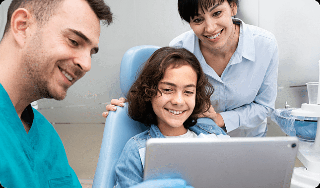 A young person, who appears to be of Middle Eastern descent based on their features and complexion, smiles with a dental professional and a woman, who may be of Asian descent based on her features, while looking at a tablet. Limestone Hills Orthodontics in Austin, TX.