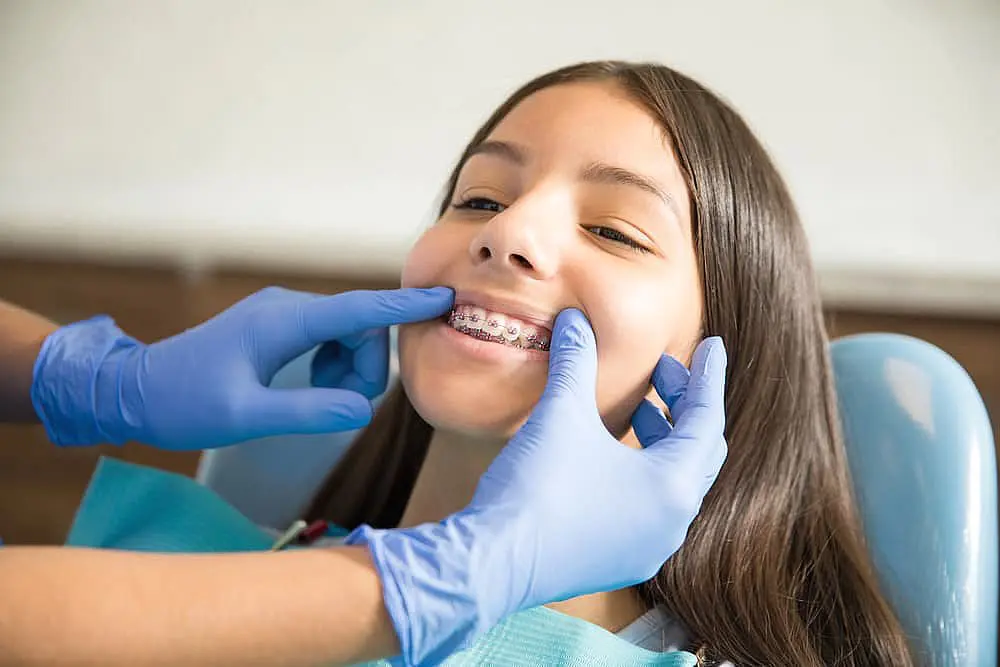 A young person, who appears to be female and may be of Hispanic or Latino descent based on their features and complexion, has their braces checked by gloved hands. Limestone Hills Orthodontics in Austin, TX.