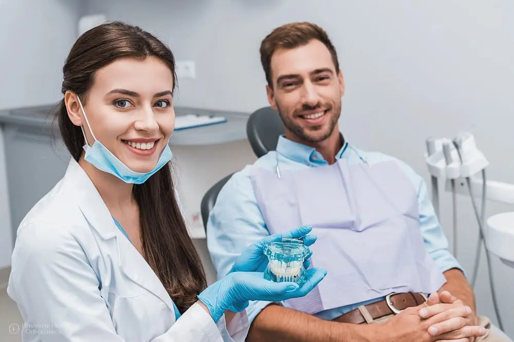 Attractive orthodontist wearing latex gloves holding dental tools near smiling patient in clinic after explaining him about overbite vs underbite