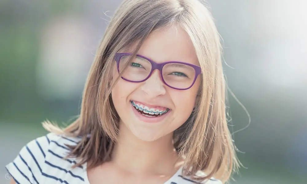 Happy teenage girl with metal braces and glasses with orthodontic rubber bands, smiling at the camera.