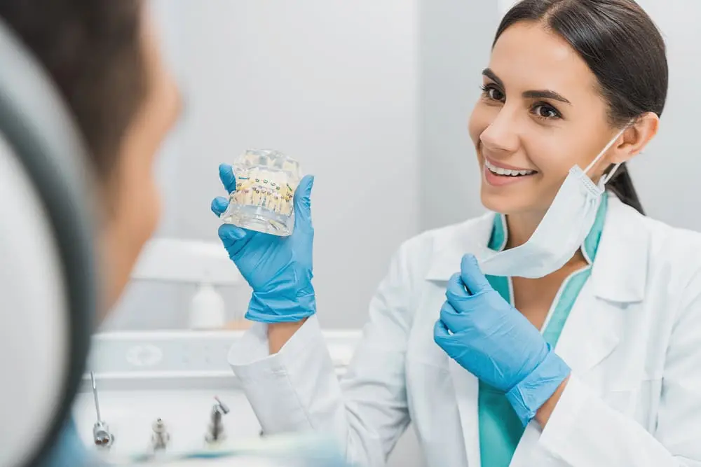 Female orthodontist smiling while showing a dental jaw model with braces to explain about the causes of crooked teeth