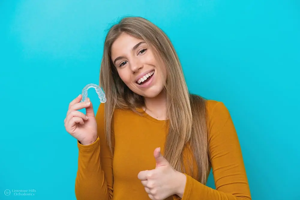 A smiling young woman, who appears to be of Caucasian descent based on her features, holds a clear aligner and gives a thumbs-up. Limestone Hills Orthodontics in Austin, TX, offers clear braces.