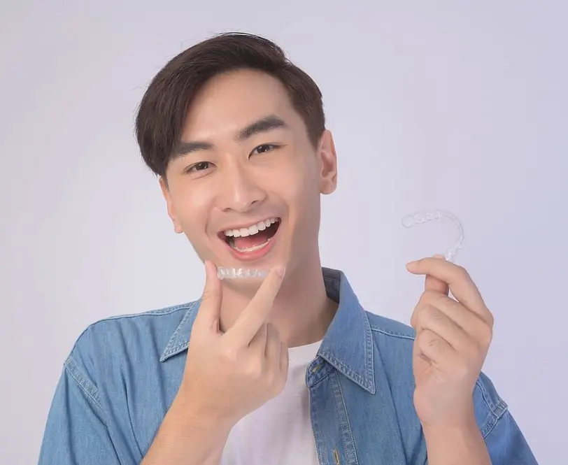 Young man in a light blue shirt smiling while holding clear aligners after not worrying about getting invisalign with missing teeth - Invisalign With Missing Teeth: A Clinical Guide | Limestone Hills Orthodontics Austin TX