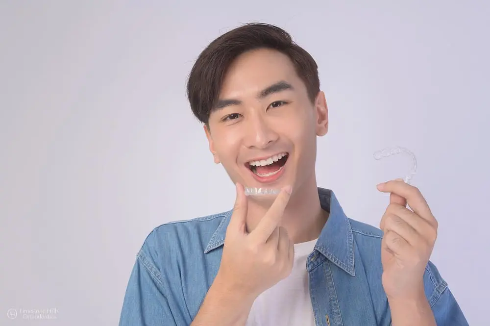 A smiling young man, who appears to be of East Asian descent based on his features, points to his teeth while holding clear aligners. Limestone Hills Orthodontics in Austin, TX, offers these.