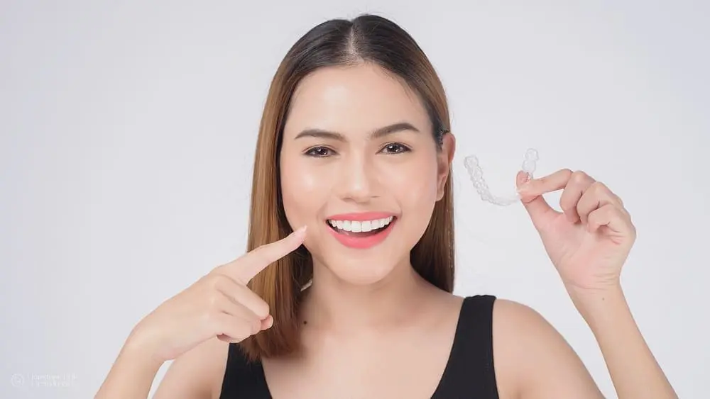 Smiling woman holding Invisalign clear aligners in a dental studio setting after wondering "do I need an orthodontist for Invisalign?"