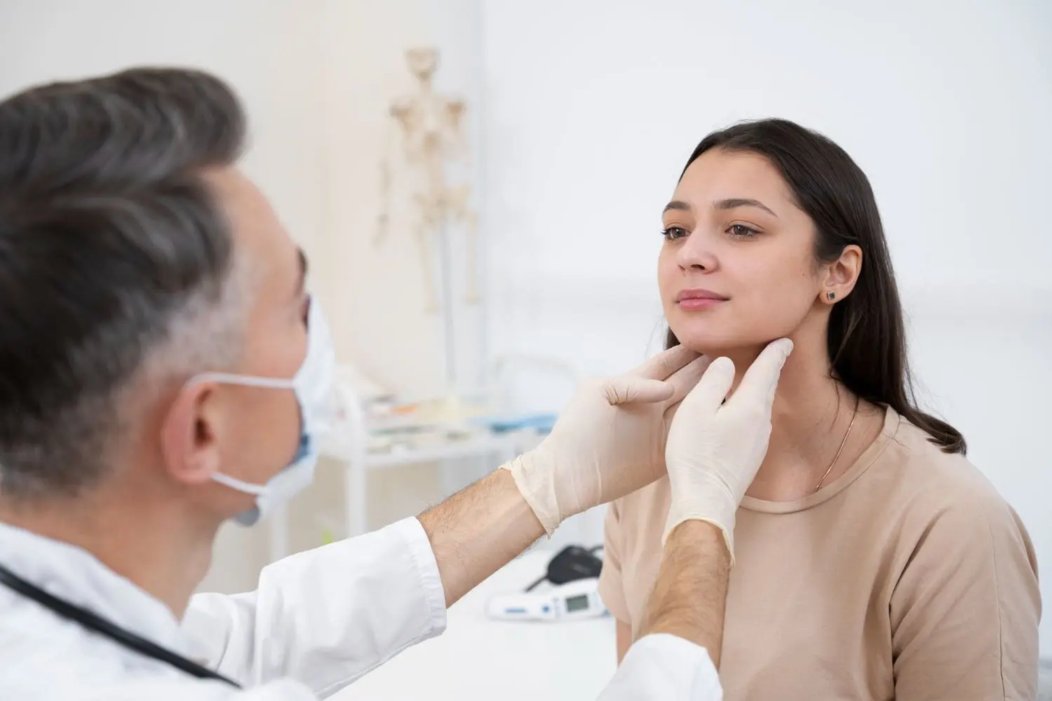 A medical professional, who appears to be a man and may be of Caucasian descent based on his visible features, wearing a face mask and gloves, is gently palpating the neck of a young woman, who appears to be of mixed ethnicity based on her features and complexion. This could be part of a general health assessment, which Limestone Hills Orthodontics in Austin, TX, might consider as part of comprehensive patient care.