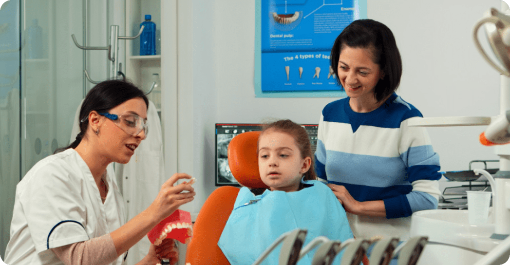 A dentist shows a dental model to a young girl sitting in a chair, while an adult stands beside them.