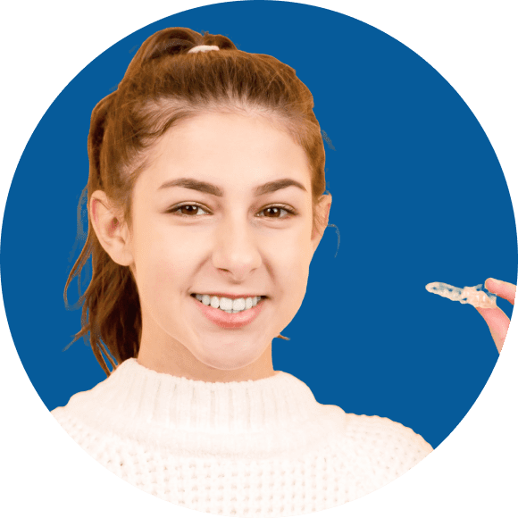 A person with long hair in a ponytail holds a clear dental aligner while smiling. The background is a solid blue.