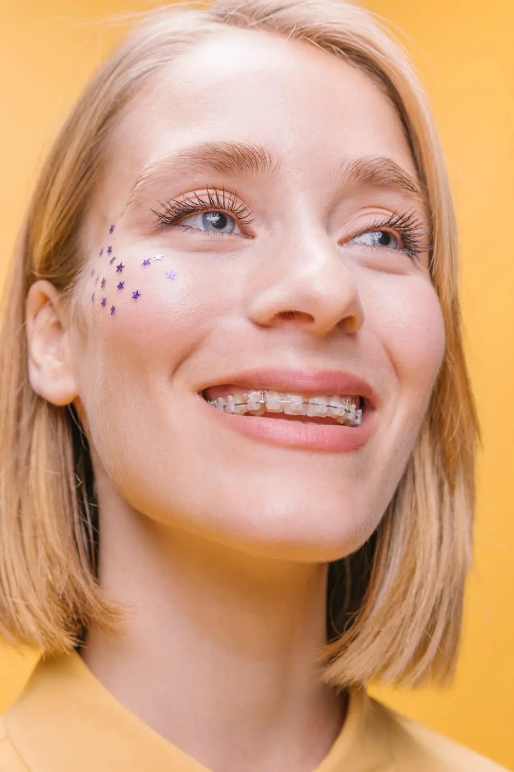 A person with ceramic braces smiles against a yellow background, small purple star stickers adorning their cheek in Austin, TX