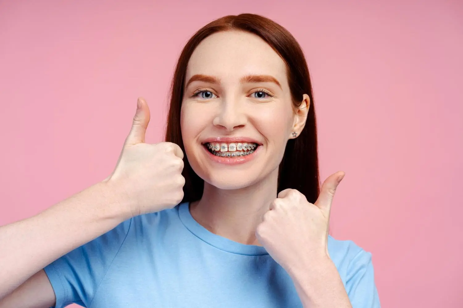 A smiling person with braces from Limestone Hills Orthodontics gives two thumbs up, proudly wearing a light blue shirt against a vibrant pink background.