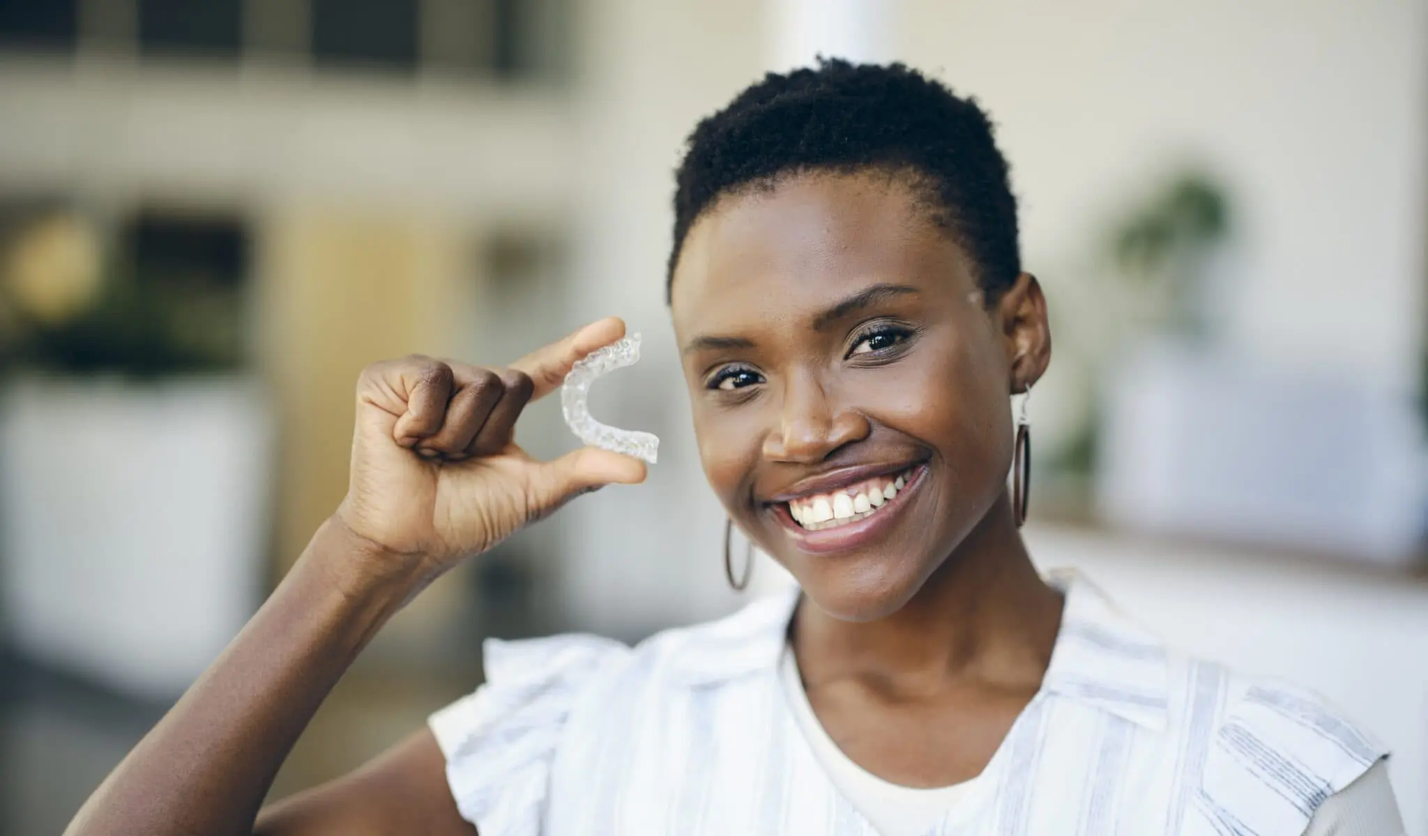 A smiling person proudly presents a clear dental aligner between their fingers, reminiscent of modern braces, in a bright room in Austin