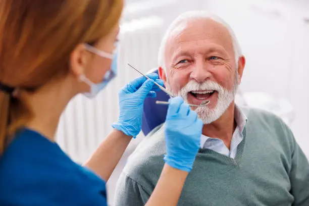 A dentist meticulously examines an older man with white hair and a beard seated in a dental chair, focusing on his oral health with the possibility of recommending jaw surgery while wearing gloves and holding dental tools in Austin