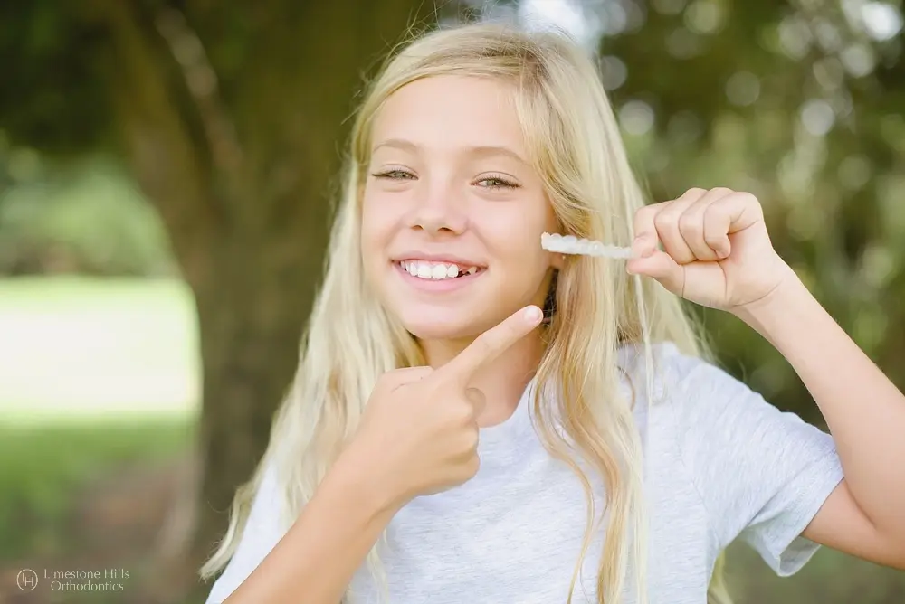Smiling blonde girl pointing at clear aligners outdoors, showing excitement about orthodontic treatment after understanding does insurance cover invisalign in Austin, TX.