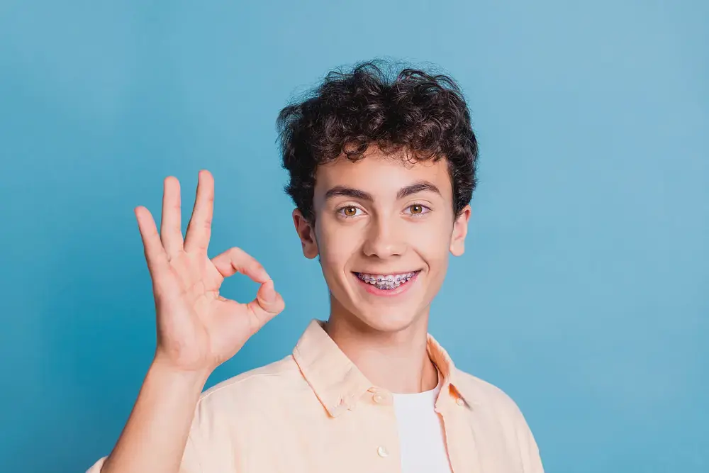 Teenage boy with braces flashes an "OK" sign, smiling for Limestone Hills Orthodontics after his tooth alignment in Austin, TX, on a blue background.