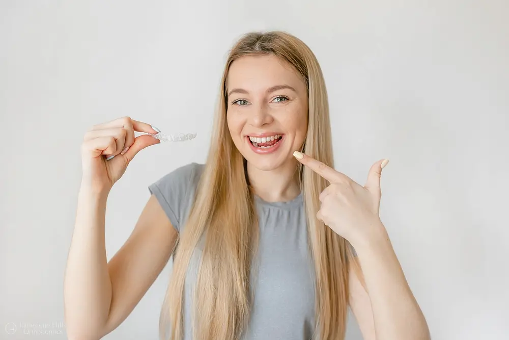 Smiling young woman with blond hair points at her teeth and holds an adult clear aligner Invisalign for Limestone Hills Orthodontics in Austin, TX.
