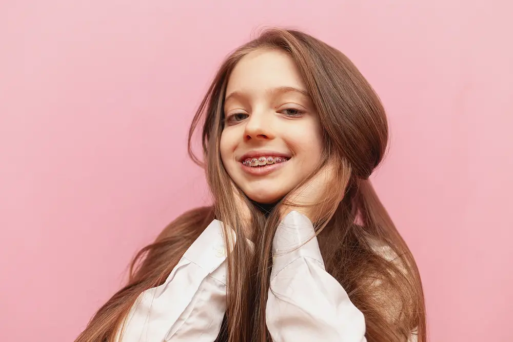 little teenage girl metal braces smiling holding long hair pink isolated background - Why Your Child Should Have Two-Phase Orthodontic Treatment in Austin, TX