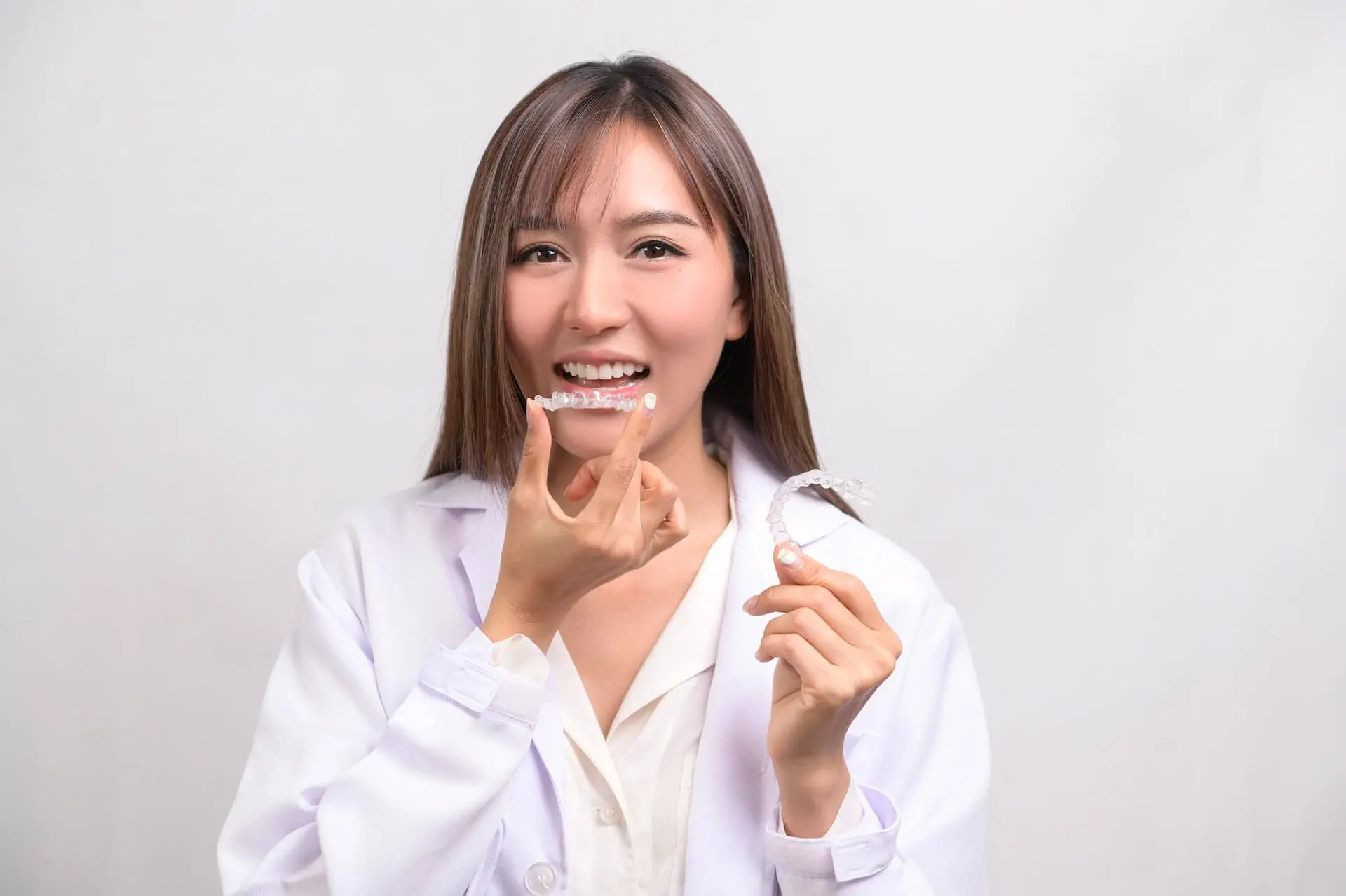 Smiling woman in a white coat holds Clear Aligners at Limestone Hills Orthodontics in Austin, TX, raising one toward her mouth.