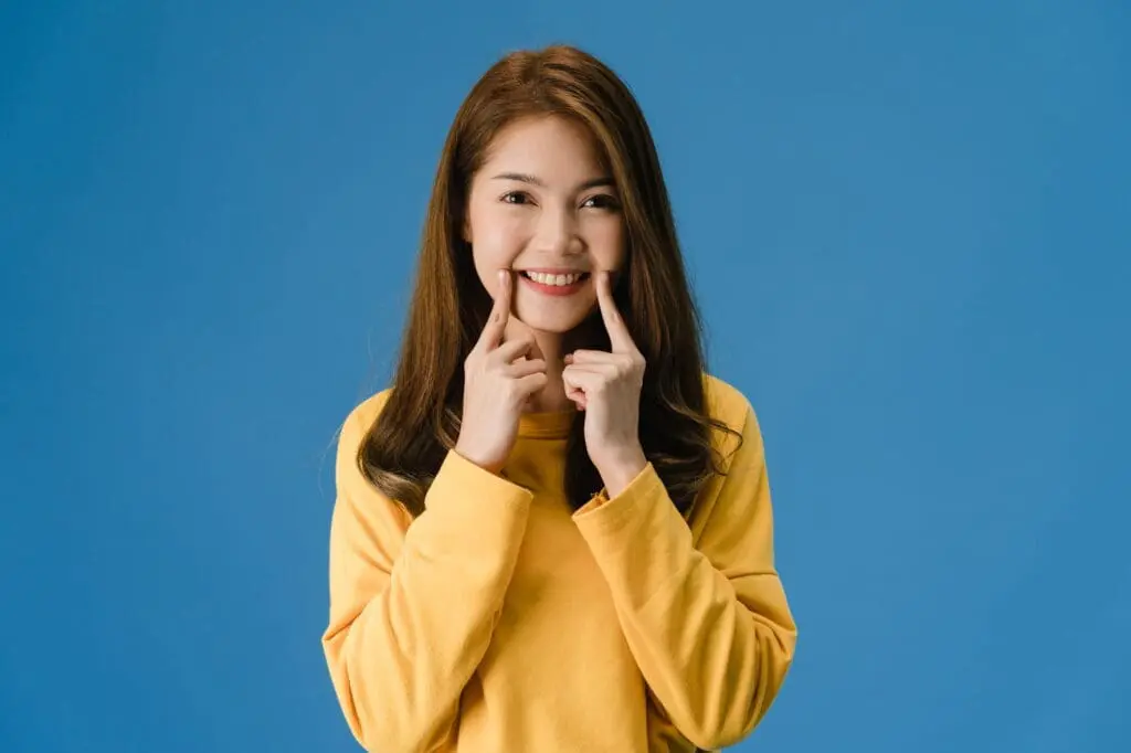 Smiling woman in yellow sweater shows off colorful braces, pointing at her smile. Limestone Hills Orthodontics in Austin, TX.