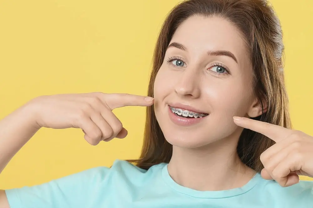 smiling woman ponting her metal braces yellow background - How Long Do Braces Take? 2026 Timeline Guide | Limestone Hills Orthodontics Austin TX