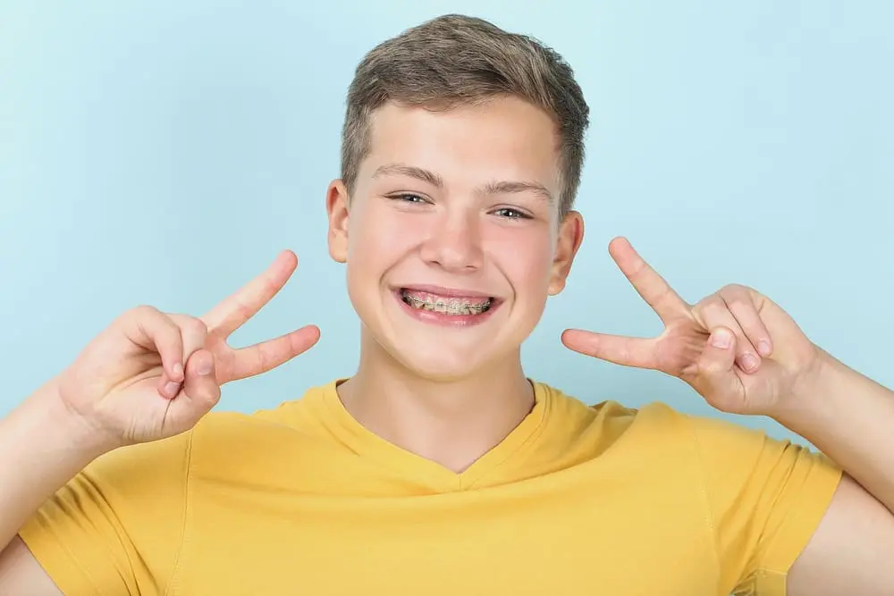 young man metal braces showing two fingers blue background - Bruxism and Orthodontics: Can Braces Help Teeth Grinding (Bruxism)? | Limestone Hills Orthodontics Austin TX