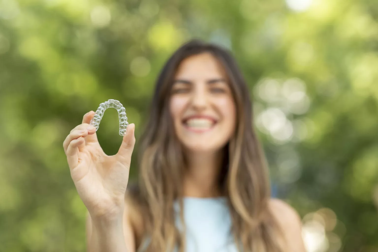 A smiling woman, who appears to be of Caucasian descent based on her features, holds a clear aligner outdoors. Limestone Hills Orthodontics in Austin, TX, offers clear braces.