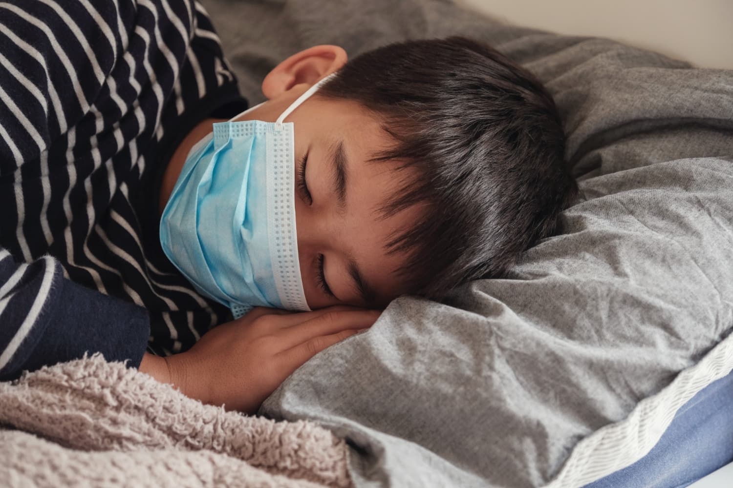 A child in a striped shirt and blue surgical mask sleeps on gray bedding at Limestone Hills Orthodontics in Austin, TX.