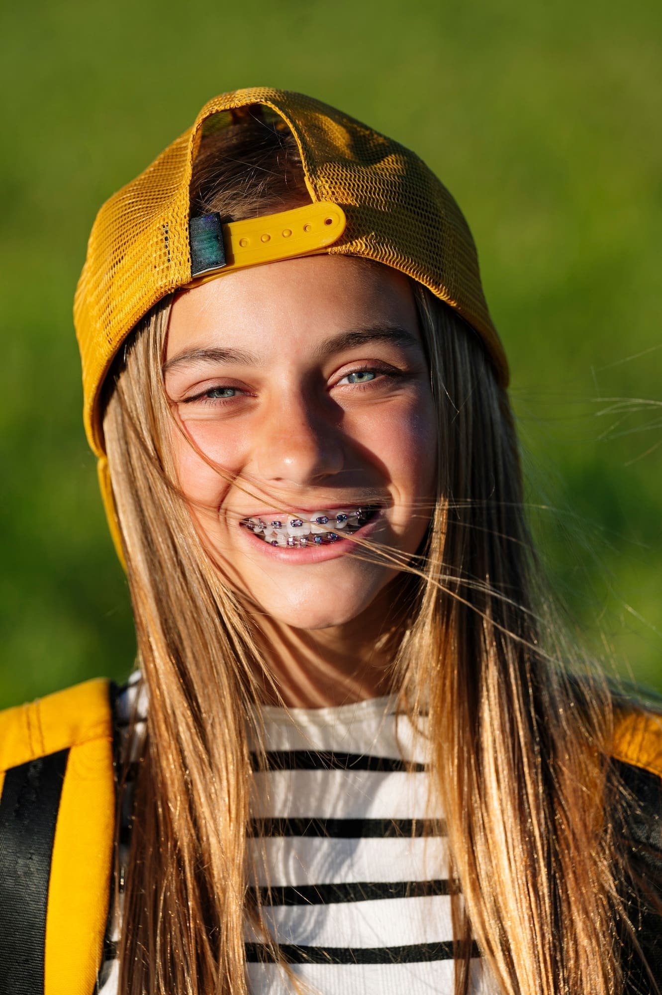 A girl with braces, long hair, and a yellow cap smiles outside on green grass for Limestone Hills Orthodontics in Austin, TX.